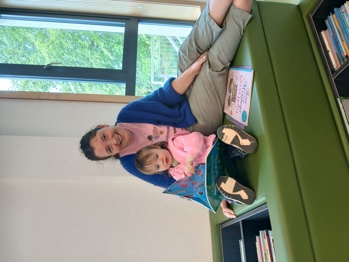 Adult and young child sitting together on a cushioned bench in a library, looking at a picture book, with additional children’s books nearby and a window in the background letting in natural light.