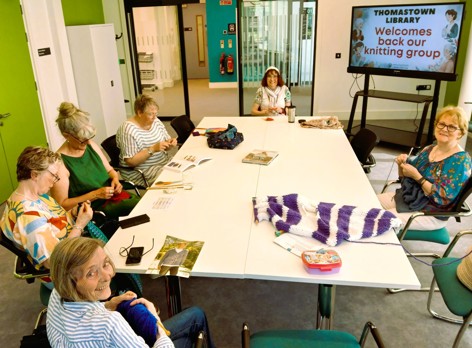Group of people seated around a table at Thomastown Library knitting together, with yarn, needles and books on the table, and a screen behind them displaying “Thomastown Library welcomes back our knitting group.”